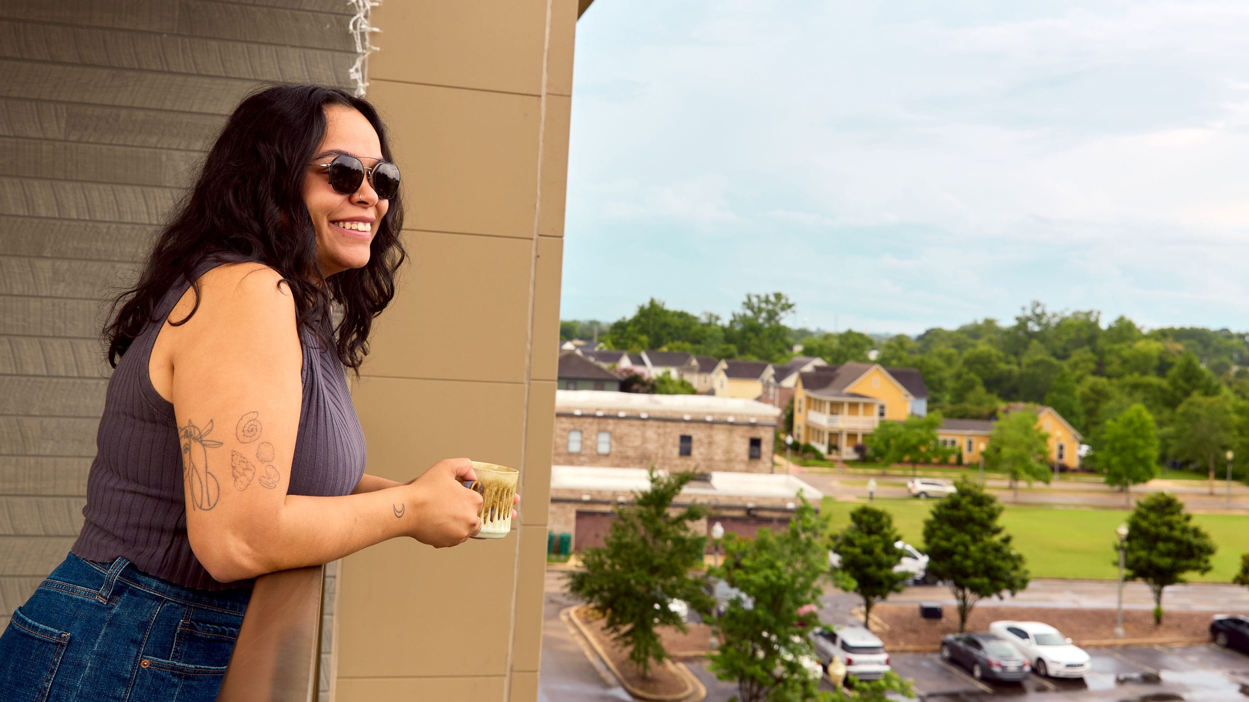 home-apartments-min woman smiling drinking coffee on apartment balcony overlooking Tupelo, MS.